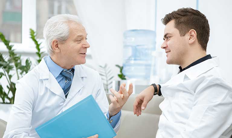 An older man in a white lab coat talks to a younger man in a white lab coat